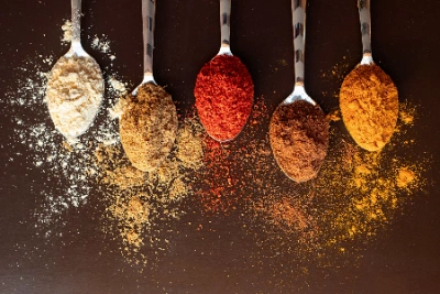 Five silver spoons filled with different colorful powdered spices like turmeric, chili, and coriander, arranged vertically on a dark brown background.