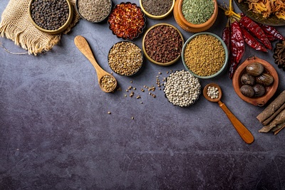 A top-down flat lay view of various whole Indian spices in small bowls and wooden spoons, arranged at the top with ample copy space below on a textured dark grey concrete background.